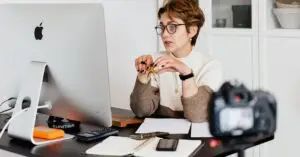A professional woman engaged in a virtual meeting setup at her home desk, using a computer and camera.